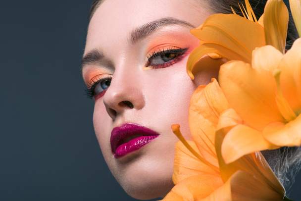 close-up portrait of attractive young woman with fashionable makeup and orange lilium flowers looking at camera isolated on grey - Foto, Imagem