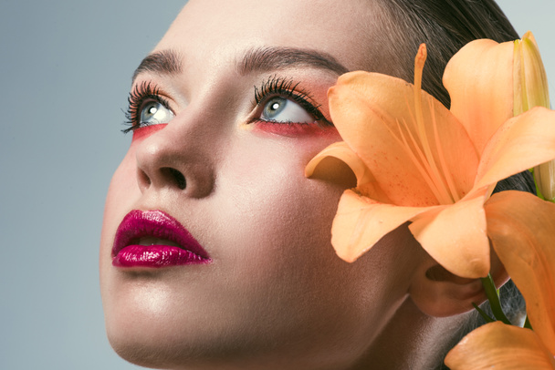 крупним планом портрет красивої молодої жінки зі стильним макіяжем і помаранчевими квітами лілії, що дивляться вгору ізольовано на сірому
 - Фото, зображення