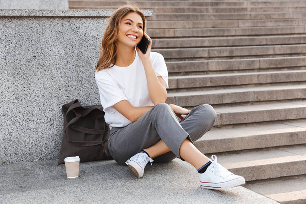Portrait of smiling young woman sitting on city stairs with legs crossed on summer day and having mobile conversation on smartphone - 写真・画像
