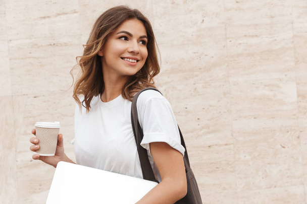 Portrait of young european woman walking against beige wall outdoor with silver laptop and takeaway coffee in hands - Photo, image