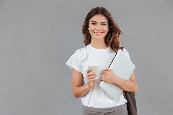 Photo of beautiful young woman smiling and standing against gray background isolated with silver laptop and takeaway coffee in hands - Фото, изображение