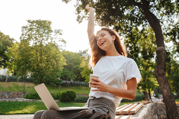Image of shappy modern woman sitting on bench in green park and enjoying summertime while using silver laptop and drinking takeaway coffee from paper cup - Foto, immagini