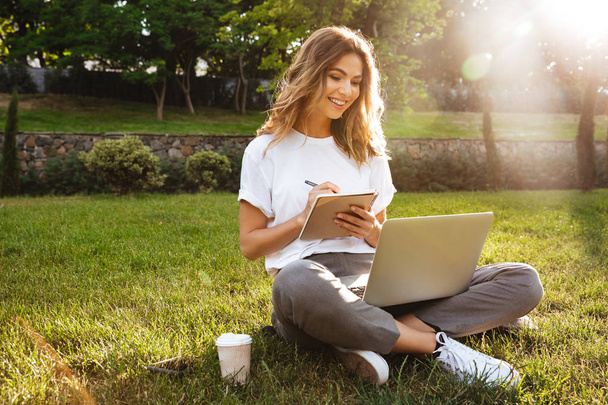 Portrait of attractive young woman sitting on green grass in park with legs crossed during summer day and writing notes with pen while using laptop - Photo, Image