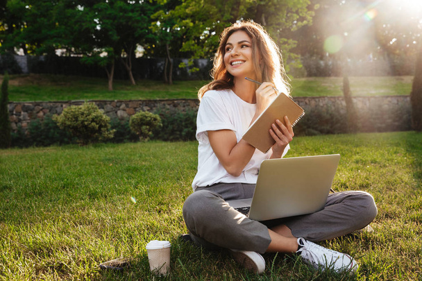 Portrait of beautiful smiling woman sitting on green grass in park with legs crossed during summer day and writing notes with pen while using laptop - Foto, afbeelding