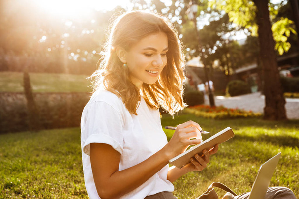 Portrait of attractive sunlit woman sitting on green grass in park during summer day and holding notebook with pen while using laptop - Valokuva, kuva