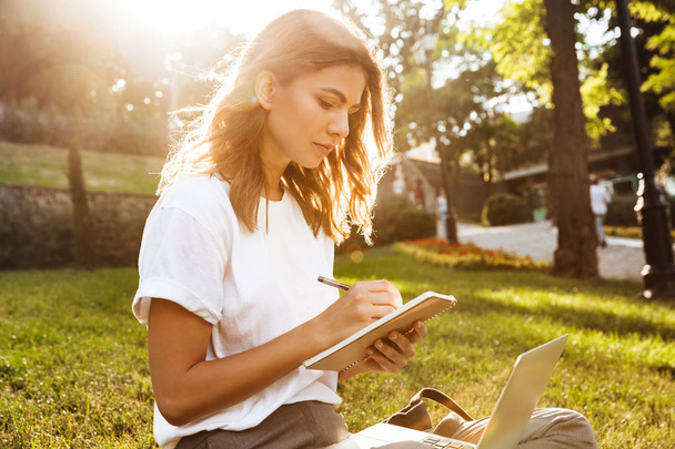Portrait of young sunlit woman sitting on green grass in park during summer day and writing notes in notebook with pen while using laptop - Fotoğraf, Görsel