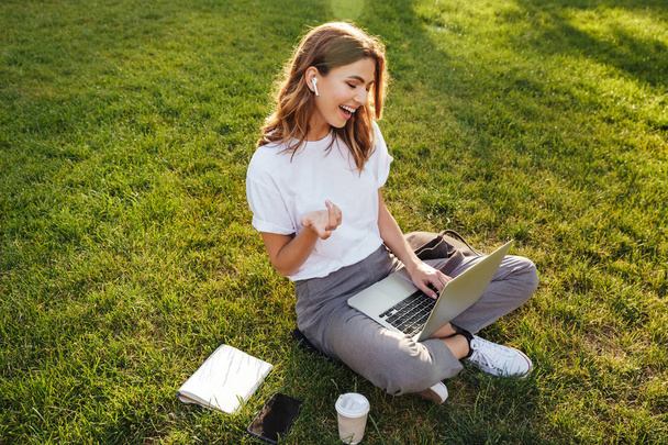 Portrait of sociable young woman sitting on green grass in park with legs crossed during summer day while using laptop and wireless earpbud for video call - Foto, Bild