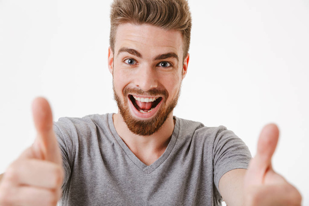 Close up portrait of a cheerful young bearded man showing thumbs up gesture isolated over white background - Фото, зображення