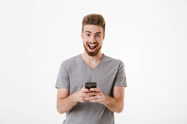 Image of happy handsome young man standing isolated over white wall background using mobile phone. Looking camera. - Φωτογραφία, εικόνα