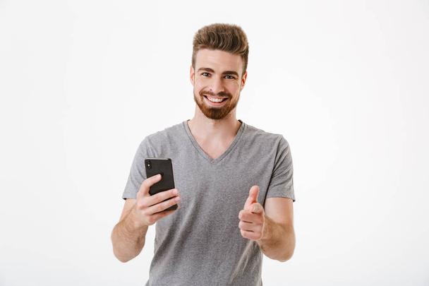 Image of happy handsome young man standing isolated over white wall background using mobile phone pointing to you. - 写真・画像