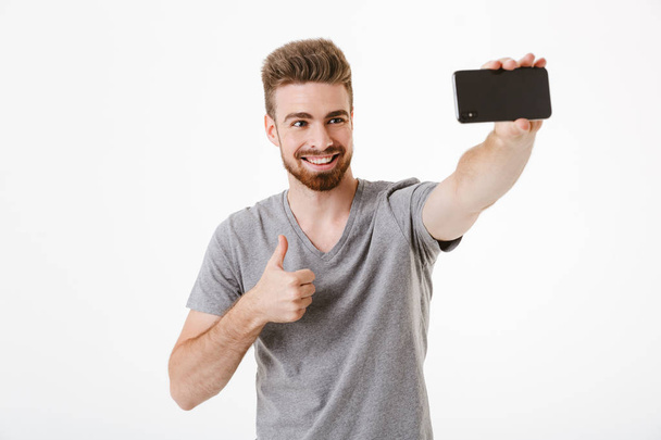 Image of happy handsome young man standing isolated over white wall background make selfie by mobile phone with thumbs up gesture. - Foto, Imagen