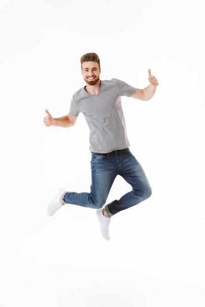 Photo of emotional excited young man jumping isolated over white wall background showing tumbs up gesture. - Fotografie, Obrázek