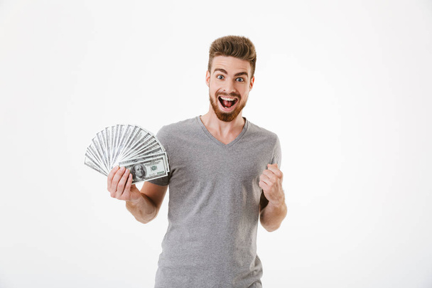 Image of excited screaming young man standing isolated over white wall background holding money make winner gesture. - Fotoğraf, Görsel