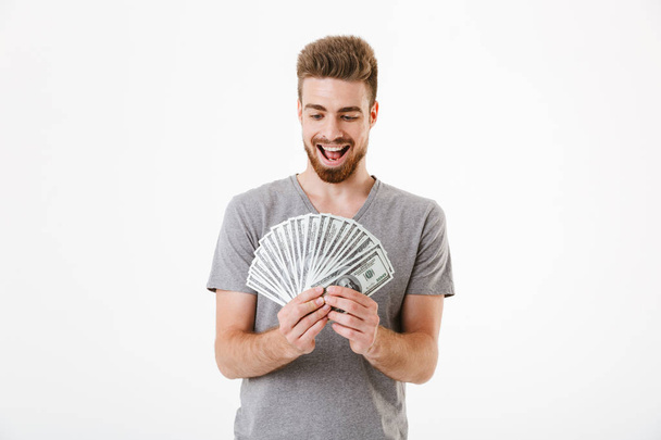 Image of excited young man standing isolated over white wall background holding money. Looking aside. - Фото, изображение