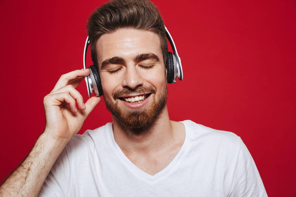 Photo of cheerful young man in white t-shirt listening music with headphones isolated over red background. - Фото, изображение