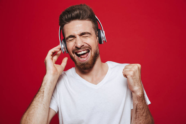 Photo of handsome emotional young man in white t-shirt listening music with headphones isolated over red background. - Valokuva, kuva