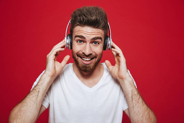 Photo of handsome emotional young man in white t-shirt listening music with headphones isolated over red background. - 写真・画像