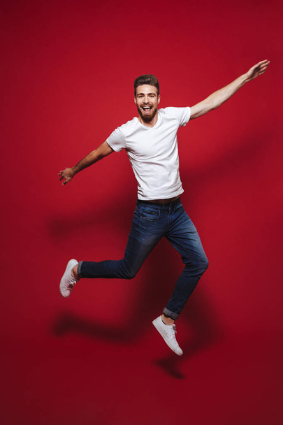 Full length portrait of a happy young bearded man celebrating success while jumping isolated over red background - Φωτογραφία, εικόνα