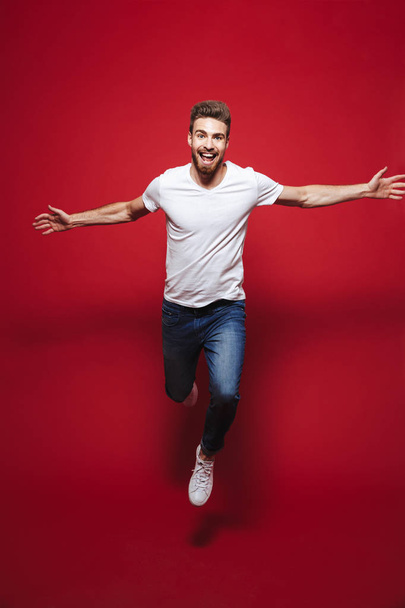 Full length portrait of a cheerful young bearded man celebrating success while jumping isolated over red background - Foto, immagini