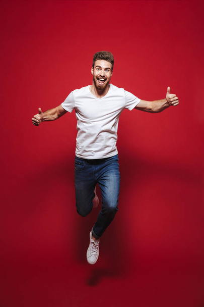 Full length portrait of a cheerful young bearded man showing thumbs up while celebrating success and jumping isolated over red background - Fotografie, Obrázek