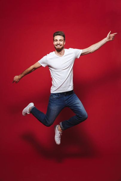 Full length portrait of a cheerful young bearded man pointing finger at copy space while celebrating success and jumping isolated over red background - Fotografie, Obrázek