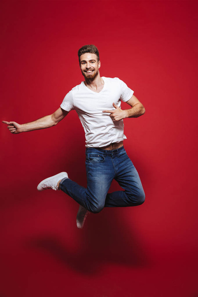 Full length portrait of a joyful young bearded man pointing finger at copy space while celebrating success and jumping isolated over red background - Фото, изображение