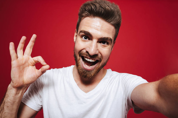 Portrait of a cheerful young bearded man showing ok gesture while taking a selfie isolated over red background - Фото, изображение