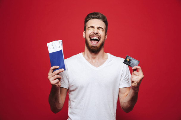 Portrait of a satisfied young bearded man holding passport with tickets and showing plastic credit card isolated over red background - Фото, изображение