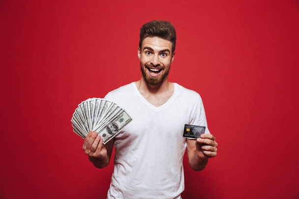 Portrait of a satisfied young bearded man holding money banknotes and showing plastic credit card isolated over red background - Photo, image