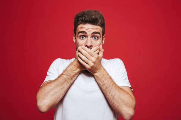 Portrait of a shocked young bearded man cover mouth with hands isolated over red background - Фото, зображення