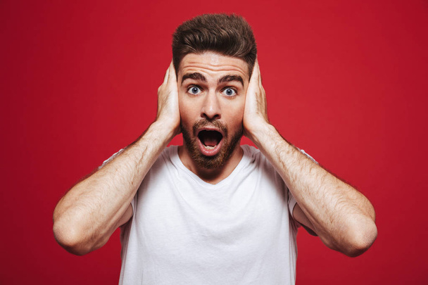 Portrait of a shocked young bearded man cover ears with hands isolated over red background - Foto, afbeelding