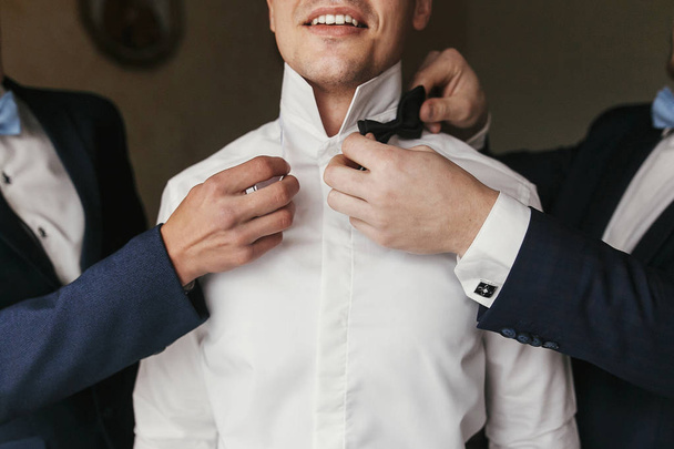 Groomsmen helping groom getting ready in the morning in the room. man hands putting on jacket and bow tie, standing at window light in hotel. preparation for wedding day - Фото, зображення