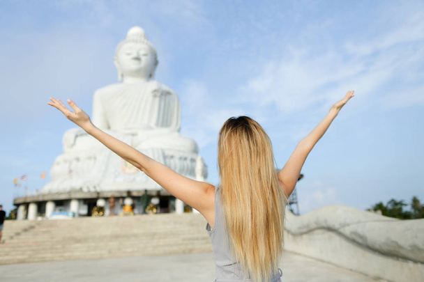 Mladá blondýnka s otevřenou náručí, stojící poblíž betonová socha Buddhy v Phuketu. - Fotografie, Obrázek