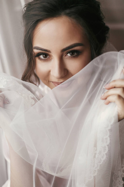 gorgeous bride portrait with make-up in the morning in room near window. beautiful woman getting ready for wedding day, holding veil and smiling. face close up. sensual moment - Foto, imagen