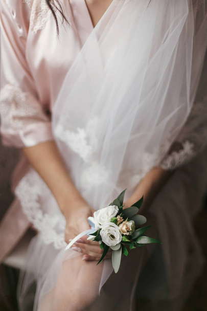 gorgeous bride holding boutonniere, sitting in robe near window. beautiful woman getting ready for wedding day, holding flowers. girl legs and veil. sensual moment. boudoir - Photo, Image