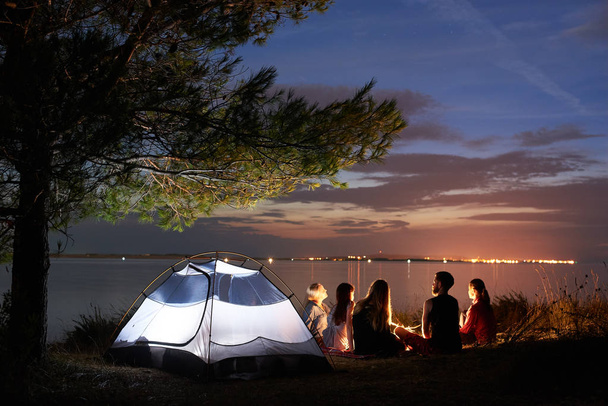 Widok z pięciu turystów, Kobieta i mężczyzna siedzi na brzegu morza wokół ogniska w namiocie pod drzewem na powierzchni gładkiej wodzie i niebieski wieczorne niebo w tle zachodu słońca z tyłu. Turystyka i camping koncepcja. - Zdjęcie, obraz