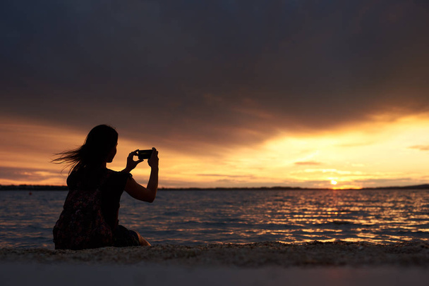 dunkle Silhouette einer Touristin mit Rucksack, die allein am Ufer am Wasserrand sitzt und das wunderschöne Meer bei Sonnenuntergang vor dunklem Abendhimmel fotografiert. Tourismus- und Urlaubskonzept. - Foto, Bild