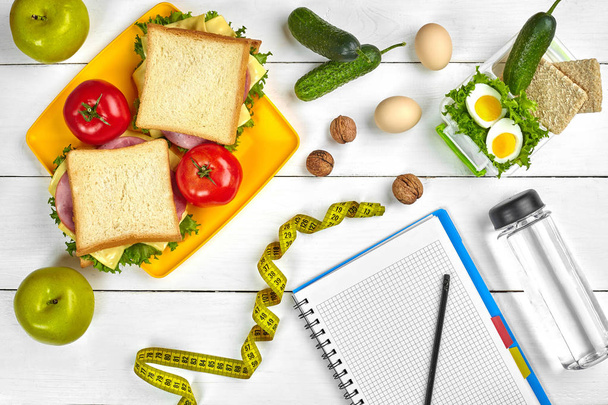 Planungsnotizbuch von oben mit Kopierraum und gesundem Frühstück. Sandwich mit Schinken und Käse, Eiern, Gurken, Tomaten und einer Flasche Wasser auf einem weißen Holztisch. - Foto, Bild