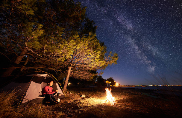 Nuit camping au bord de la mer. Femme randonneuse préparant la nourriture sur un brûleur à gaz, se reposant à l'entrée de la tente touristique au feu de camp près de la forêt contre un ciel étoilé bleu vif avec la Voie lactée
 - Photo, image