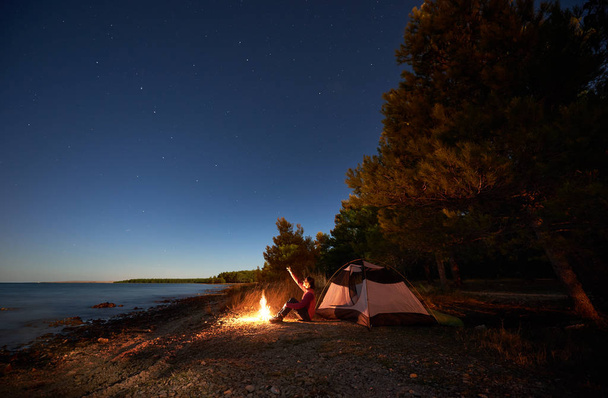 Night camping at sea shore. Woman hiker sitting in front of tourist tent at campfire, pointing to starry sky, enjoying beautiful view of clear blue water. Tourism and active lifestyle concept - Photo, Image