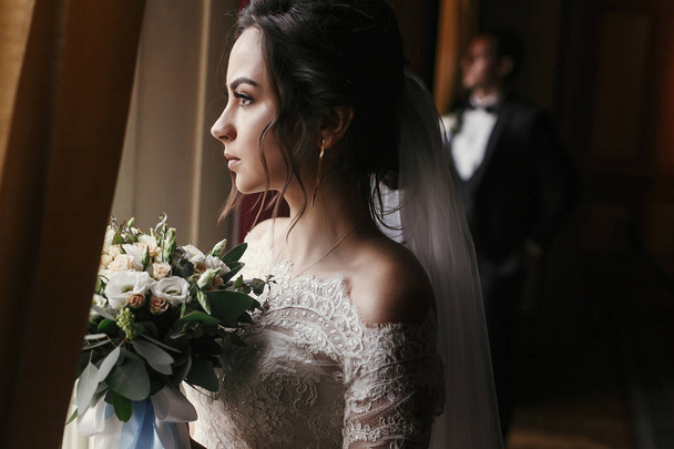 великолепная невеста в изумительном платье с букетом и стильным женихом, позирующим у окна в роскошном номере отеля. богатая супружеская пара смотрит на свет. романтический момент молодоженов в классическом помещении
 - Фото, изображение