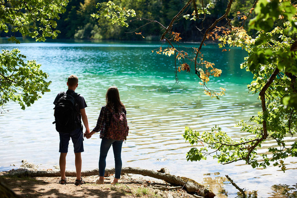 Back view of tourist couple backpacks standing on river bank holding hands enjoying beautiful colorful spring panorama of lit by sun green forest and blue lake. - Foto, Imagen