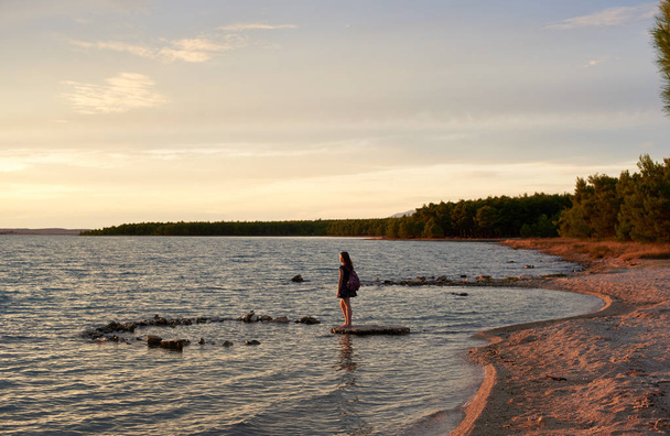 Jonge vrouw in korte jurk en met kleurrijke rugzak staan blote voeten op vlakke steen in ondiep water, niet ver van de kust op rustige avond. Toerisme en vakanties concept. - Foto, afbeelding