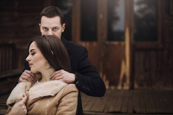nádherná nevěsta v kabátě a stylový ženicha pózuje v dřevěném domku v zimním lese. šťastný svatební pár jemně objímá v zasněženém parku. stodola svatba. smyslné romantické chvíle - Fotografie, Obrázek