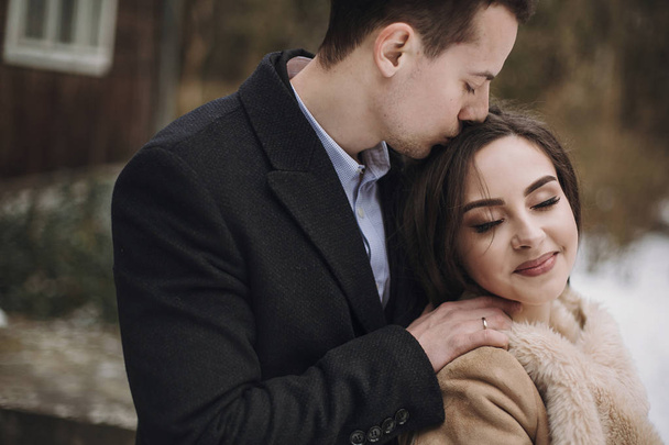 gorgeous bride in coat and stylish groom hugging at wooden house. wedding couple gently hugging and embracing in snowy park. barn wedding. romantic sensual moment of newlyweds - Фото, изображение