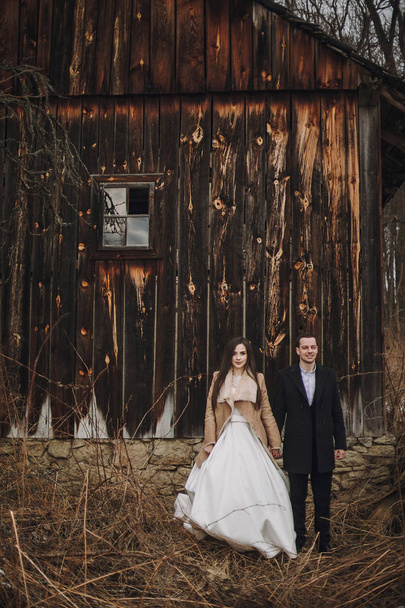 gorgeous bride in coat and stylish groom posing at wooden house. wedding couple gently hugging and embracing in snowy park. barn wedding. romantic sensual moment - Фото, зображення
