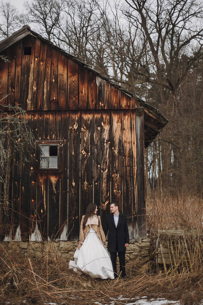 πανέμορφη νύφη σε παλτό και κομψό γαμπρό που ποζάρουν στο ξύλινο σπίτι. ζευγάρι γάμος απαλά Αγκαλιάζοντας και Αγκαλιάζοντας στο χιονισμένο πάρκο. Γάμος αχυρώνα. Ρομαντικά αισθησιακό παρόν - Φωτογραφία, εικόνα