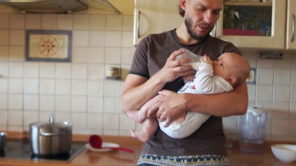 Stylish multitasking father feeds the baby in the kitchen and boils the soup, stirring it on the stove. A man has a stylish hairstyle and a modern beard - Video