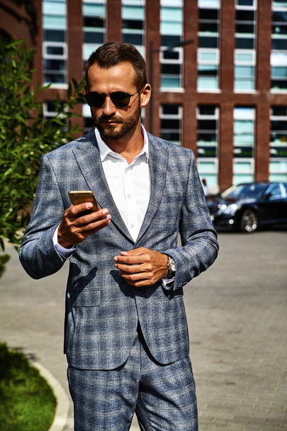Portrait of sexy handsome fashion businessman model dressed in elegant checkered suit having business mobile conversation on smartphone on street. Metrosexual - Фото, зображення