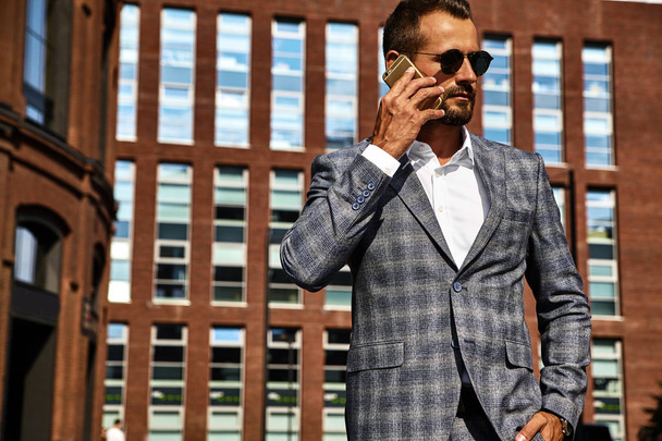 Portrait of sexy handsome fashion businessman model dressed in elegant checkered suit having business mobile conversation on smartphone on street. Metrosexual - Zdjęcie, obraz